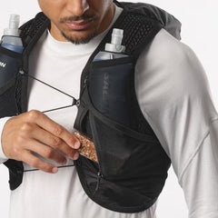 Person wearing a black hydration vest with water bottles and snacks, holding a snack.