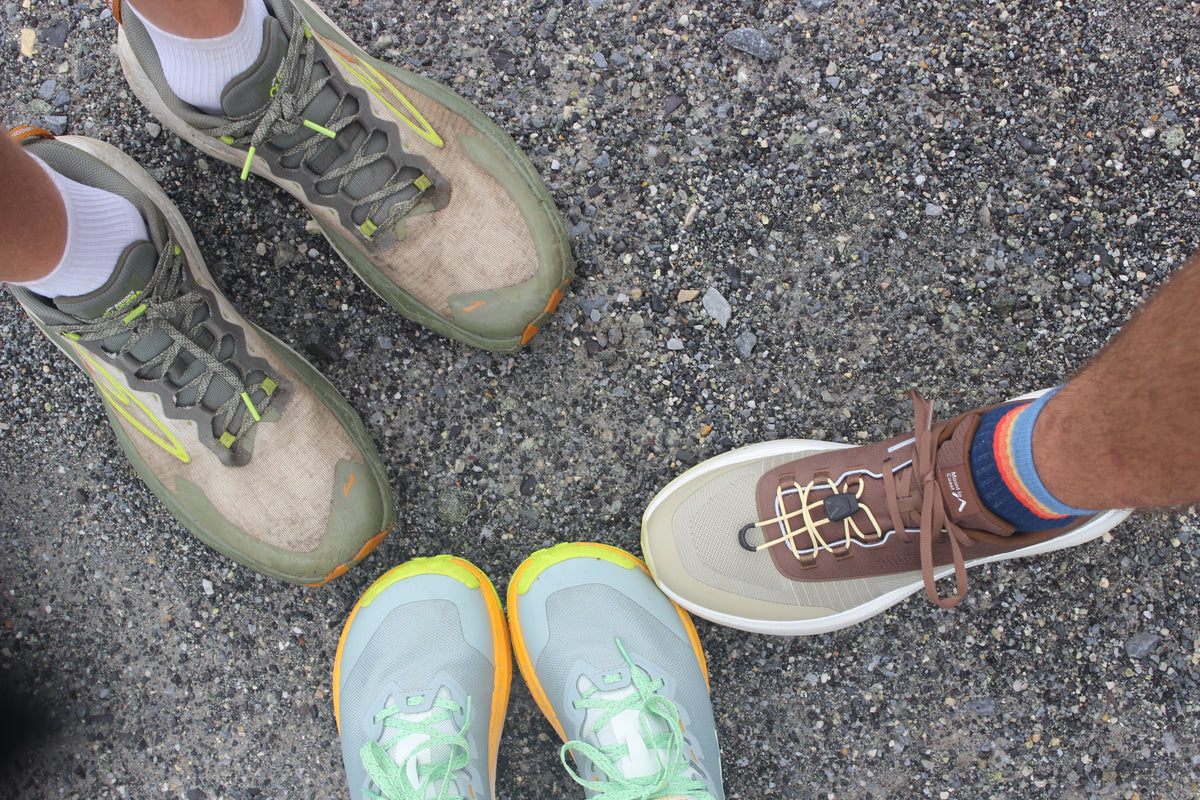 Three pairs of running shoes, including Brooks and Mount to Coast, toe to toe on gravel